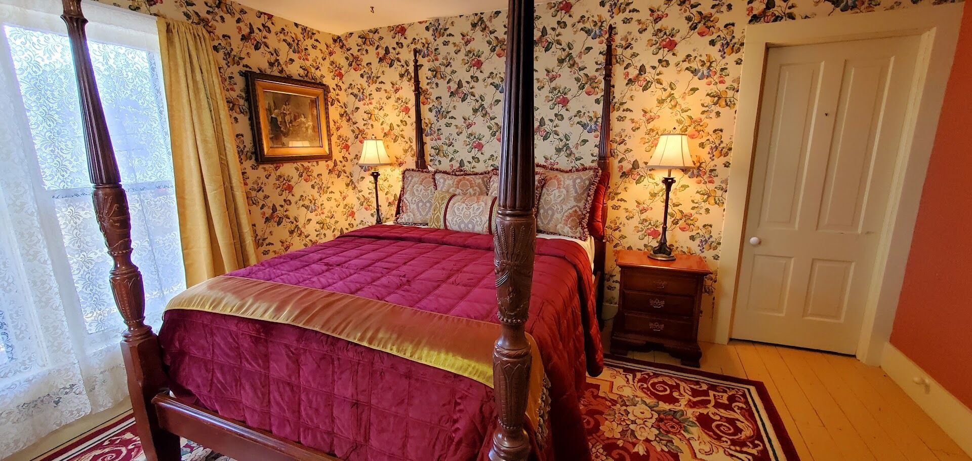 A four-poster bed with a red bedspread, two bedside tables with lamps, floral wallpaper, and a door.