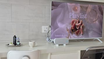 A modern kitchen with a flat-screen TV displaying a playlist, a white chair, a small table with a cup, and a decorative bottle.