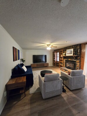 A living room with a flat-screen TV, a ceiling fan, a stone fireplace, and a bookshelf.