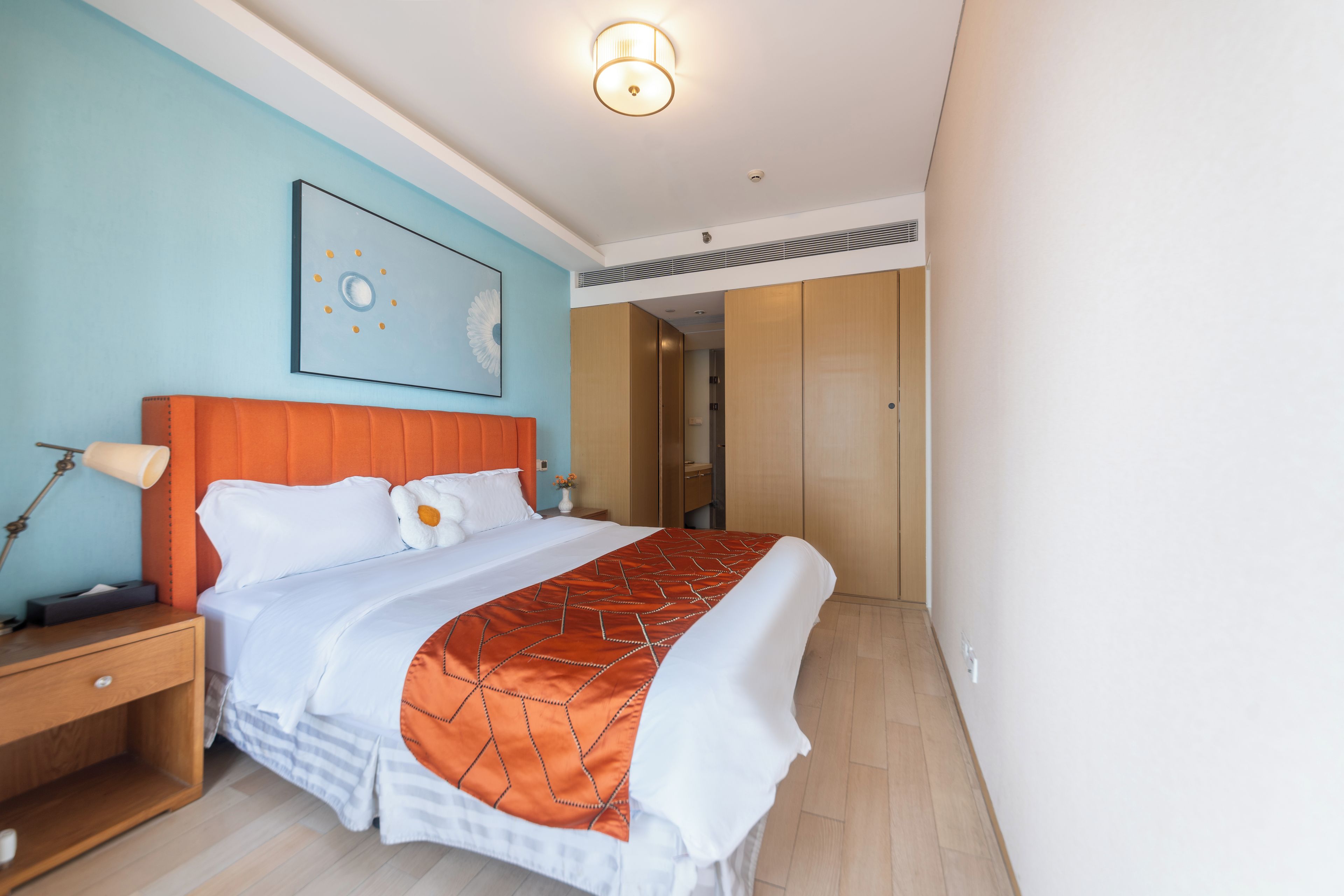 A hotel room with a bed, a nightstand, a wall-mounted lamp, and a large window.