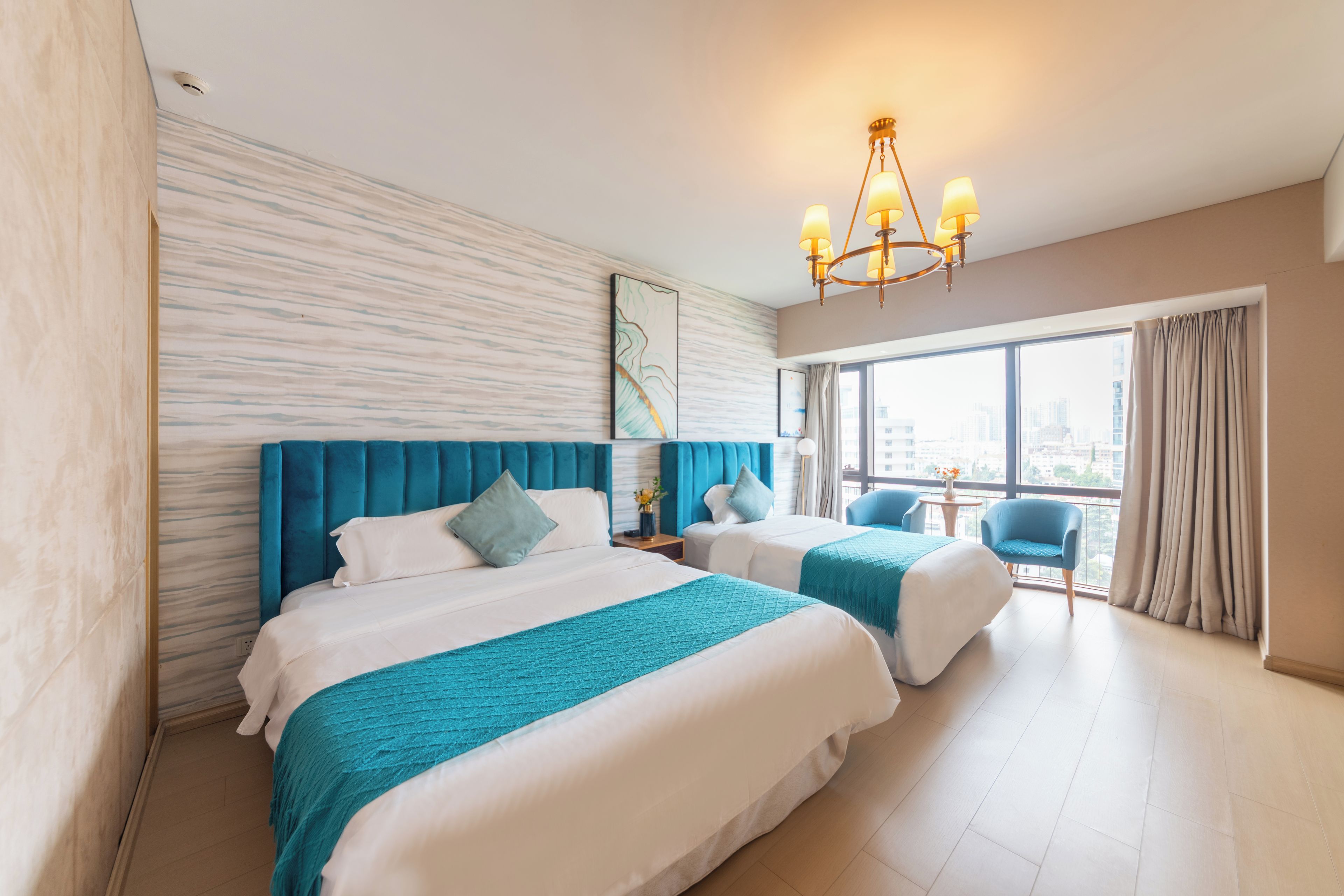 A hotel room with two beds, a large window with curtains, a city view, and a chandelier.