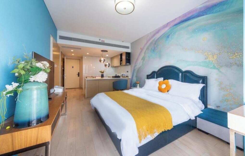 Een hotelkamer met een bed, een nachtkastje, een vaas met bloemen, een groot abstract wanddecor en uitzicht op de keuken.