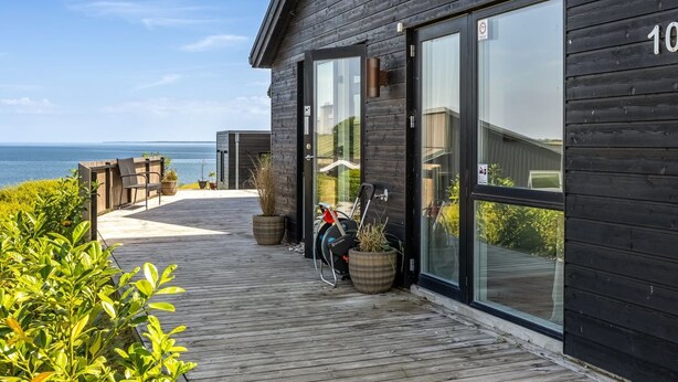 En modern byggnad i trä med stora glasdörrar, en träveranda och utsikt över havet.