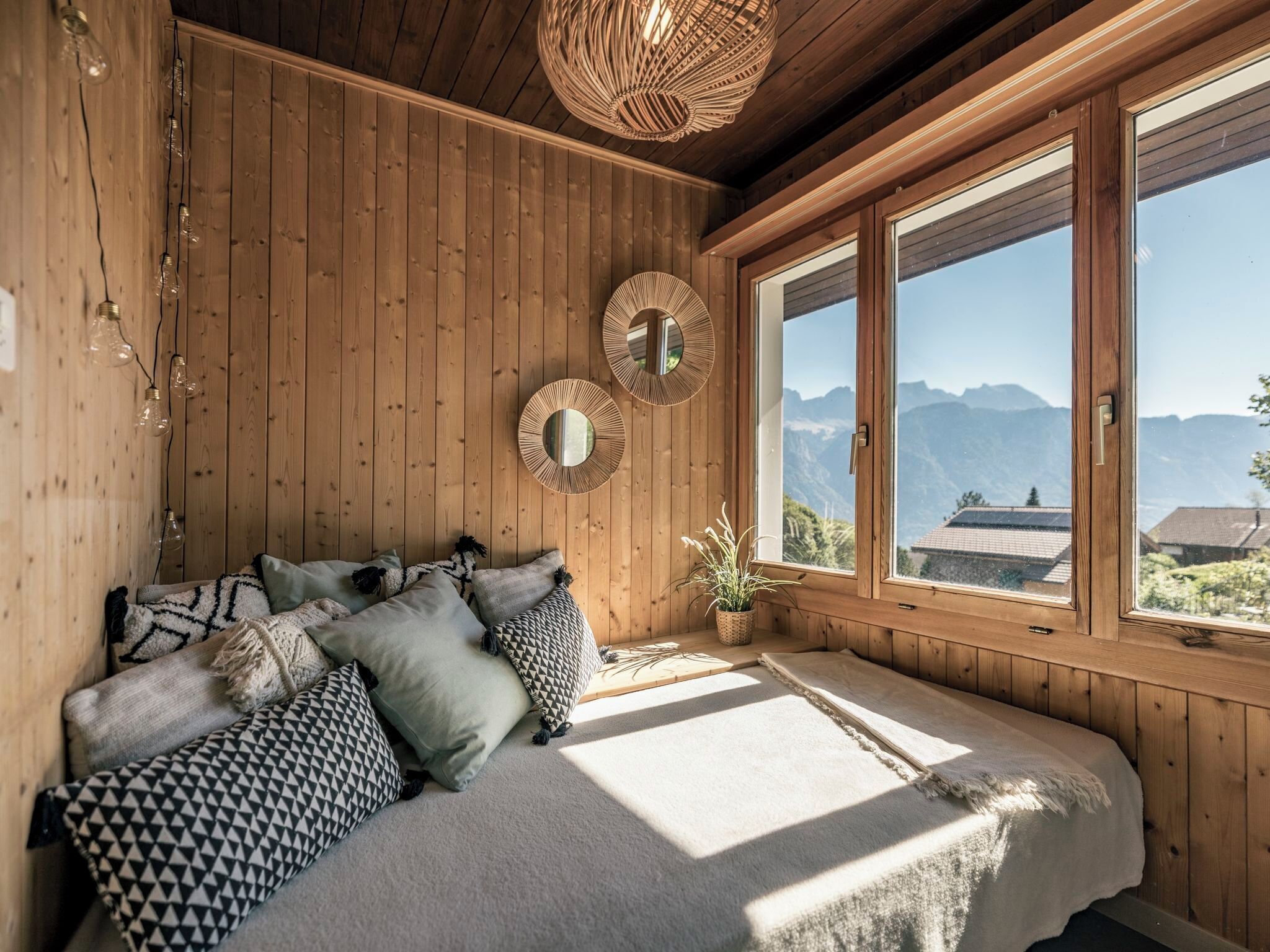 Une pièce en bois avec un lit, deux miroirs muraux et une vue sur les montagnes, visible à travers de grandes fenêtres.