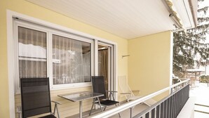 Ein Balkon mit zwei Stühlen, einem Tisch, einem Fenster mit Vorhängen und einem Geländer.