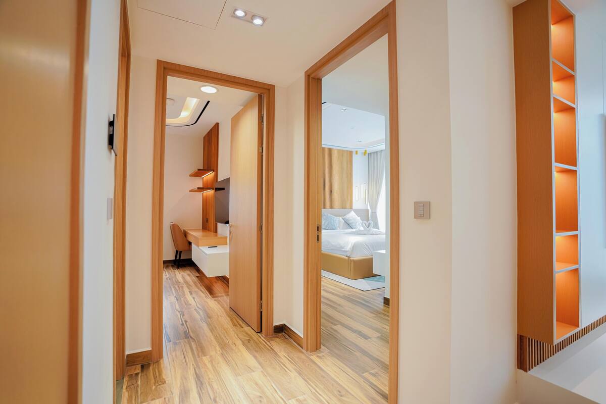 A hotel corridor with wooden flooring, a bed with a headboard, and a seating area with a chair and table.