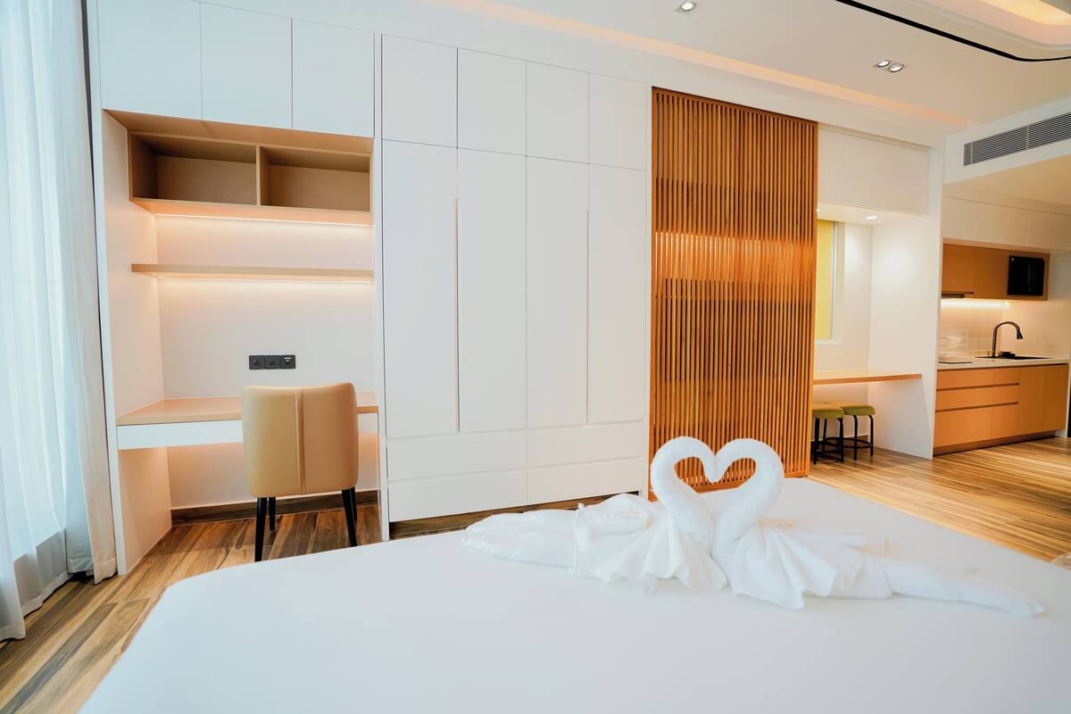 A modern hotel room with a bed, a desk, a chair, a wooden partition, and a kitchenette.