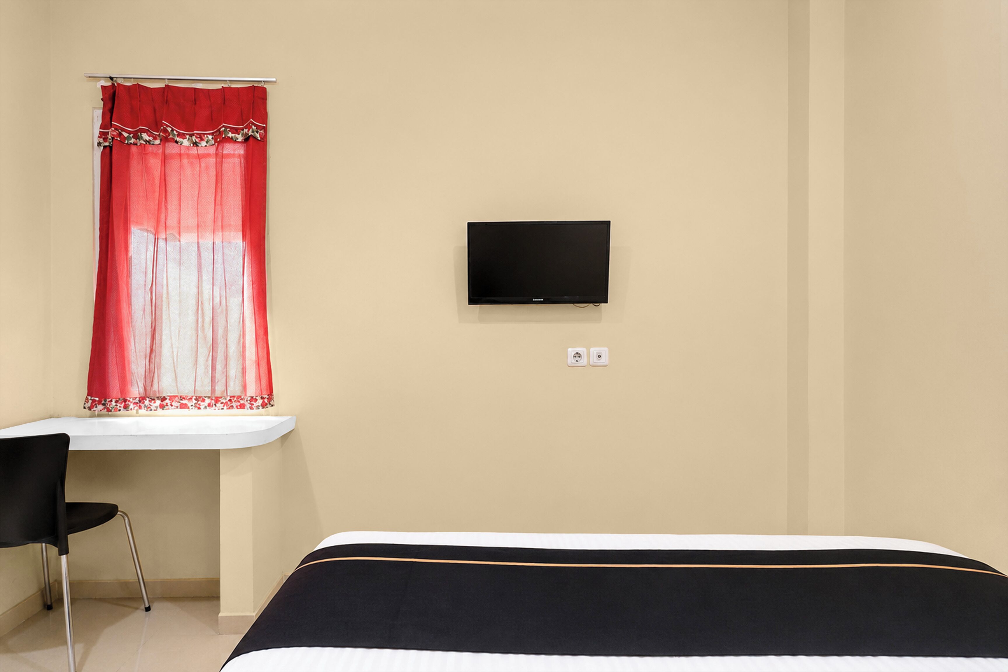 A room with a bed, a desk with a chair, a television mounted on the wall, and a window with a red curtain.