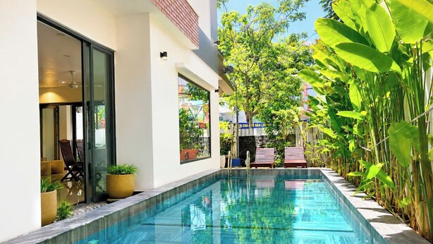 Una piscina rectangular con agua cristalina, rodeada de plantas en macetas y una entrada con puerta de cristal.