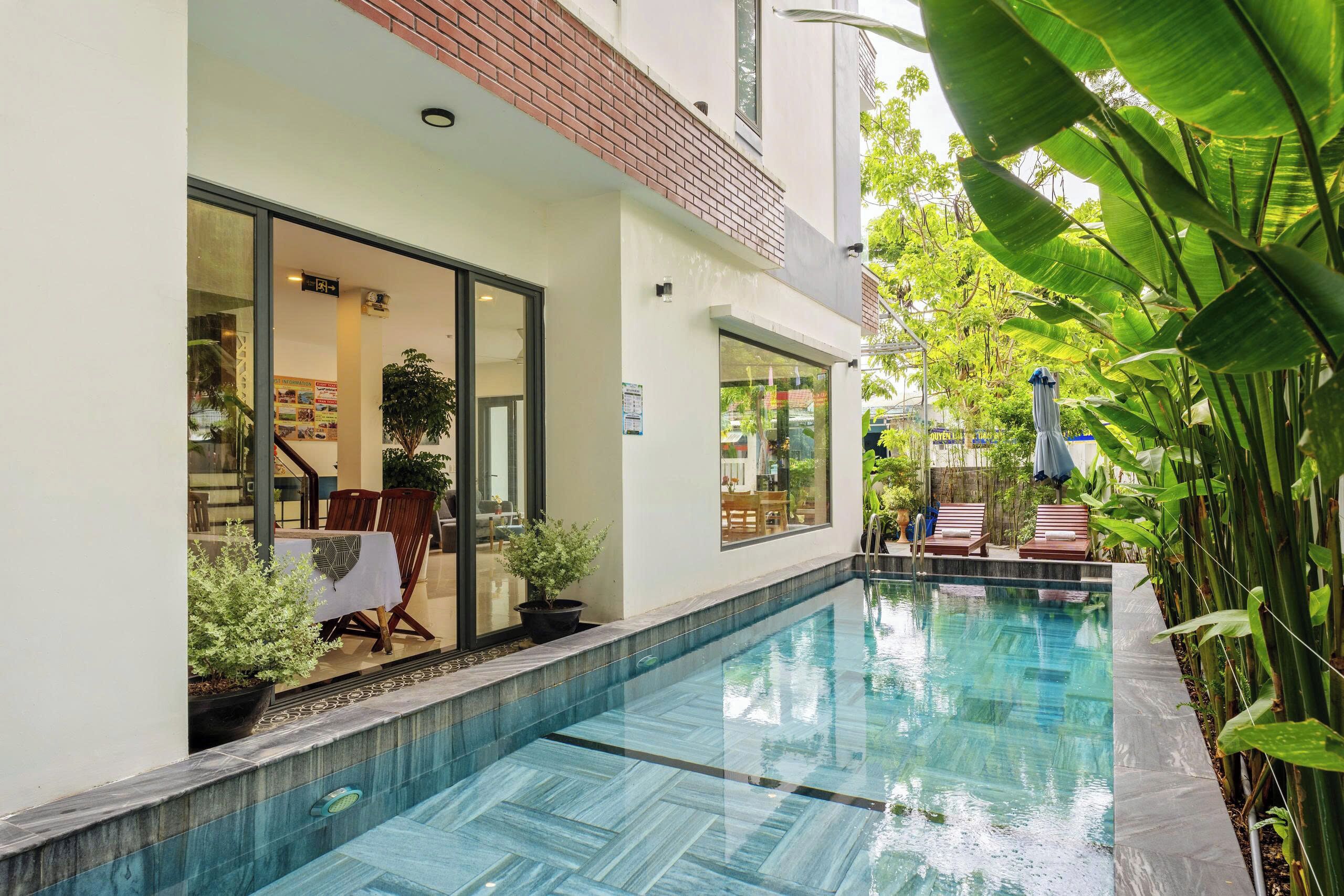Una piscina con borde de azulejos, la entrada de un edificio con puertas de vidrio y plantas en macetas.