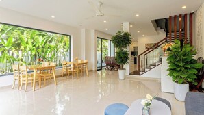 Amplia sala de estar con una mesa de comedor, sillas, una escalera y ventanas grandes con follaje verde en el exterior.
