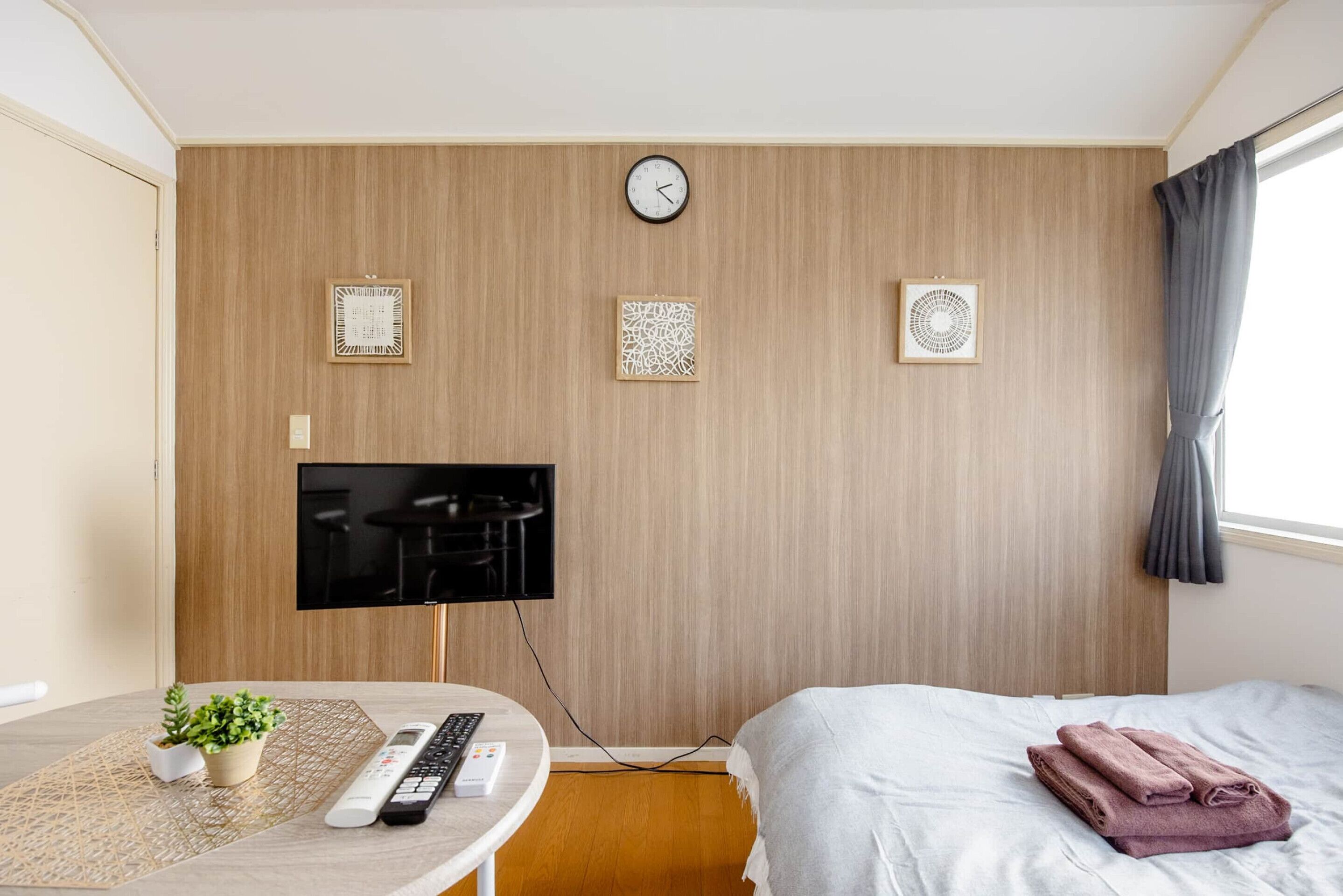 A hotel room with a bed, a TV mounted on the wall, a clock, and three framed wall art pieces.