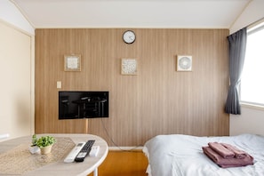 A hotel room with a bed, a TV mounted on the wall, a clock, and three framed wall art pieces. - Maison Shin-Nakano (Honcho, Nakano-ku)