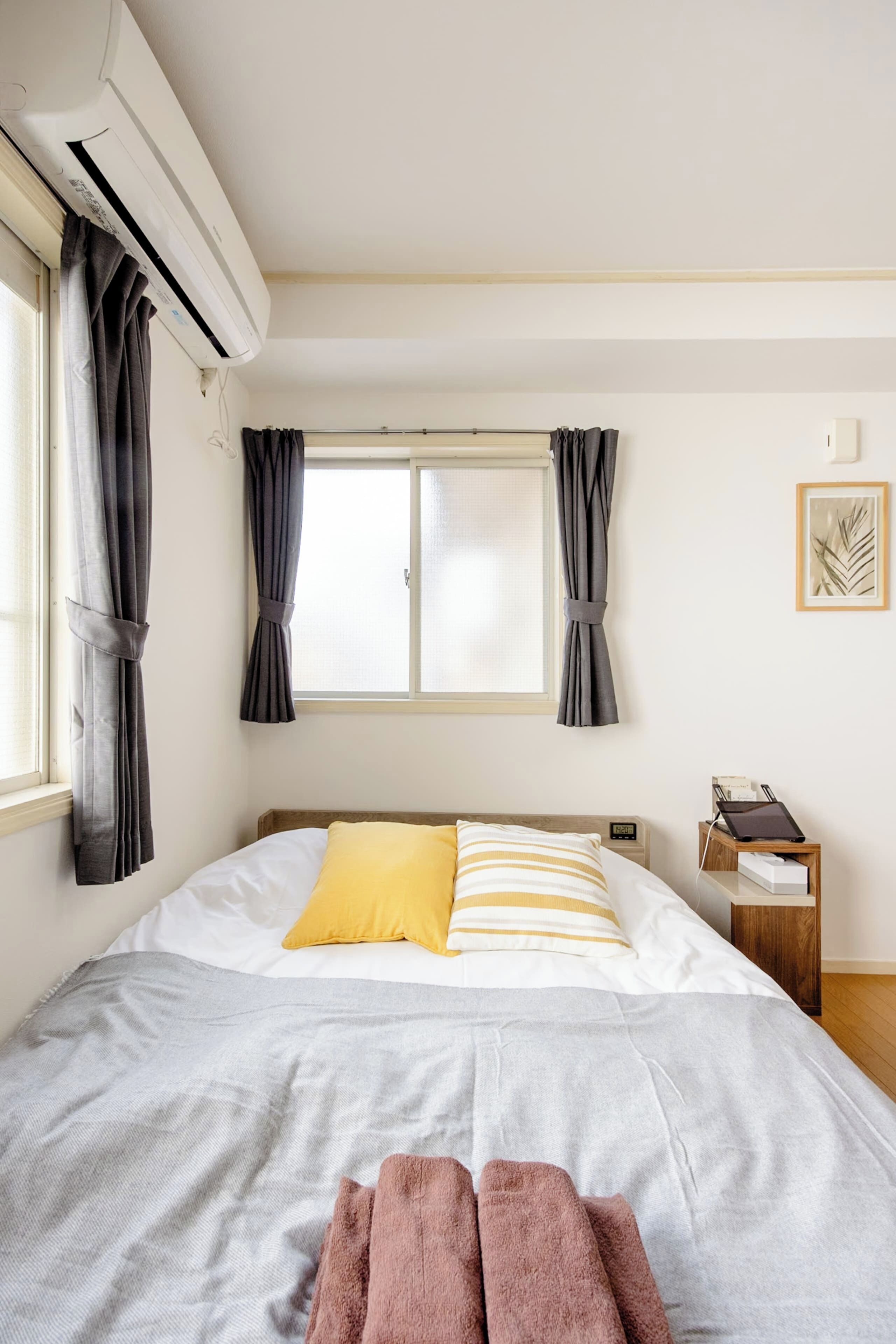 A bedroom with a bed, curtains, an air conditioner, a bedside table with a lamp, and a framed picture on the wall.