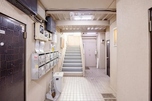 Un couloir étroit avec un escalier menant à une porte, une pièce utilitaire avec des casiers et une poubelle.