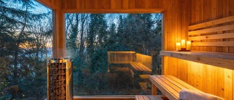 A wooden sauna room with a bench, a sauna heater, and a view of a forest.