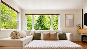 Ein modernes Wohnzimmer mit einem Ecksofa, einem großen Fenster mit Blick auf Bäume und einem flachbildfernseher an der Wand.