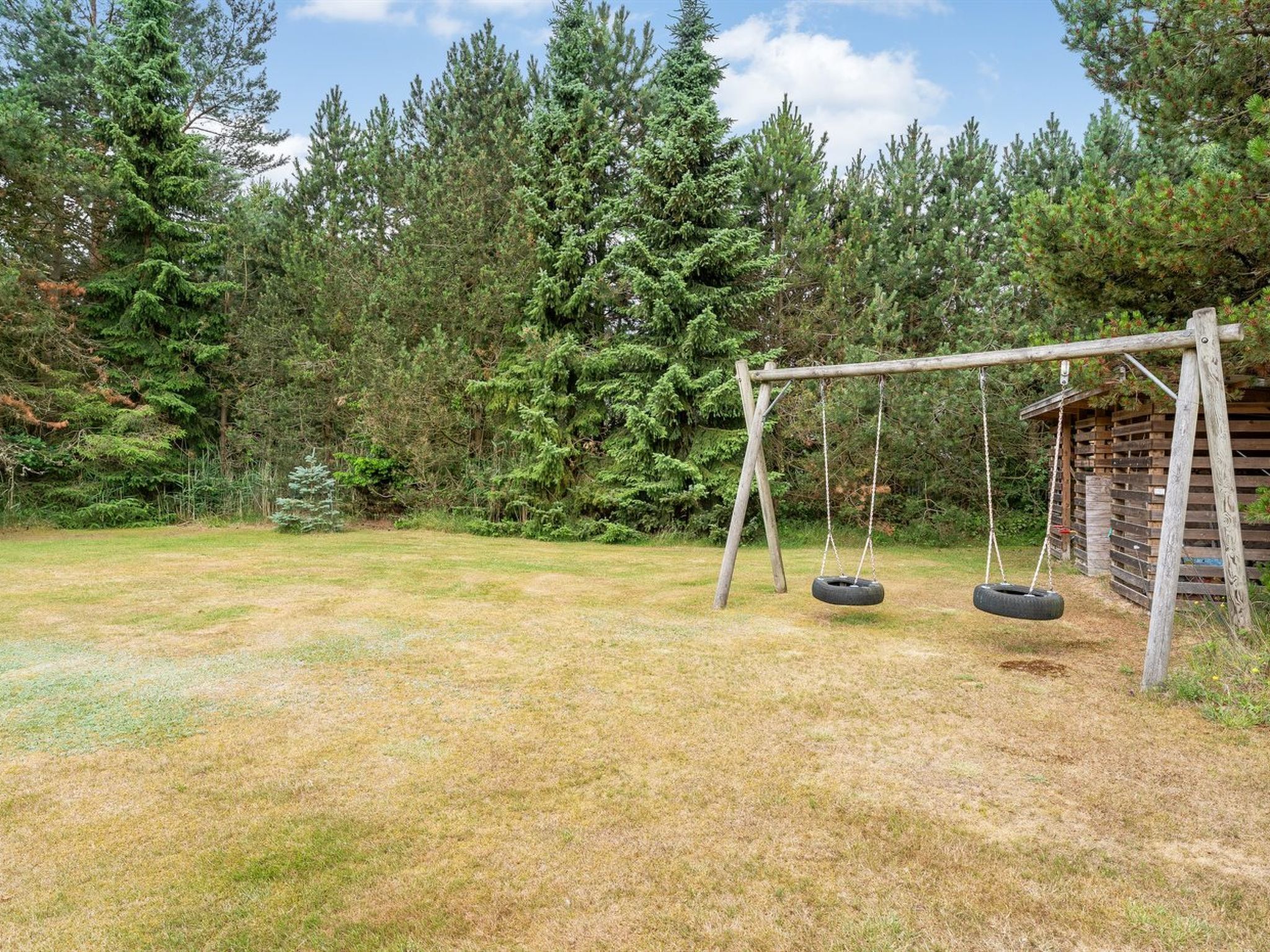 Un ensemble de jeux d’extérieur en bois, comprenant deux balançoires en forme de pneu, installé dans une cour verdoyante, avec un chalet en bois en arrière-plan.