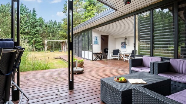 En moderne udendørs terrasse med fletmøbler, en gyngestativ og udsigt til et skovområde.