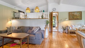 En hyggelig stue med sofa, sofabord og spisebord. Der er et køkkenområde med skabe og vask. Værelset har trægulv og et loft med synlige bjælker.