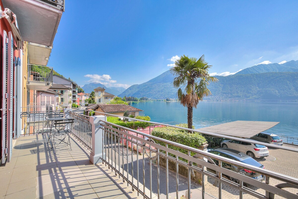 A balcony with a view of a lake and mountains, a parked car, and a palm tree.
