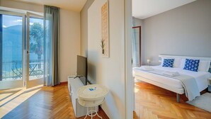 A hotel room with a bed, a desk, a chair, a TV, and a balcony with a view of the mountains. - 76 The Lake House (Melide)