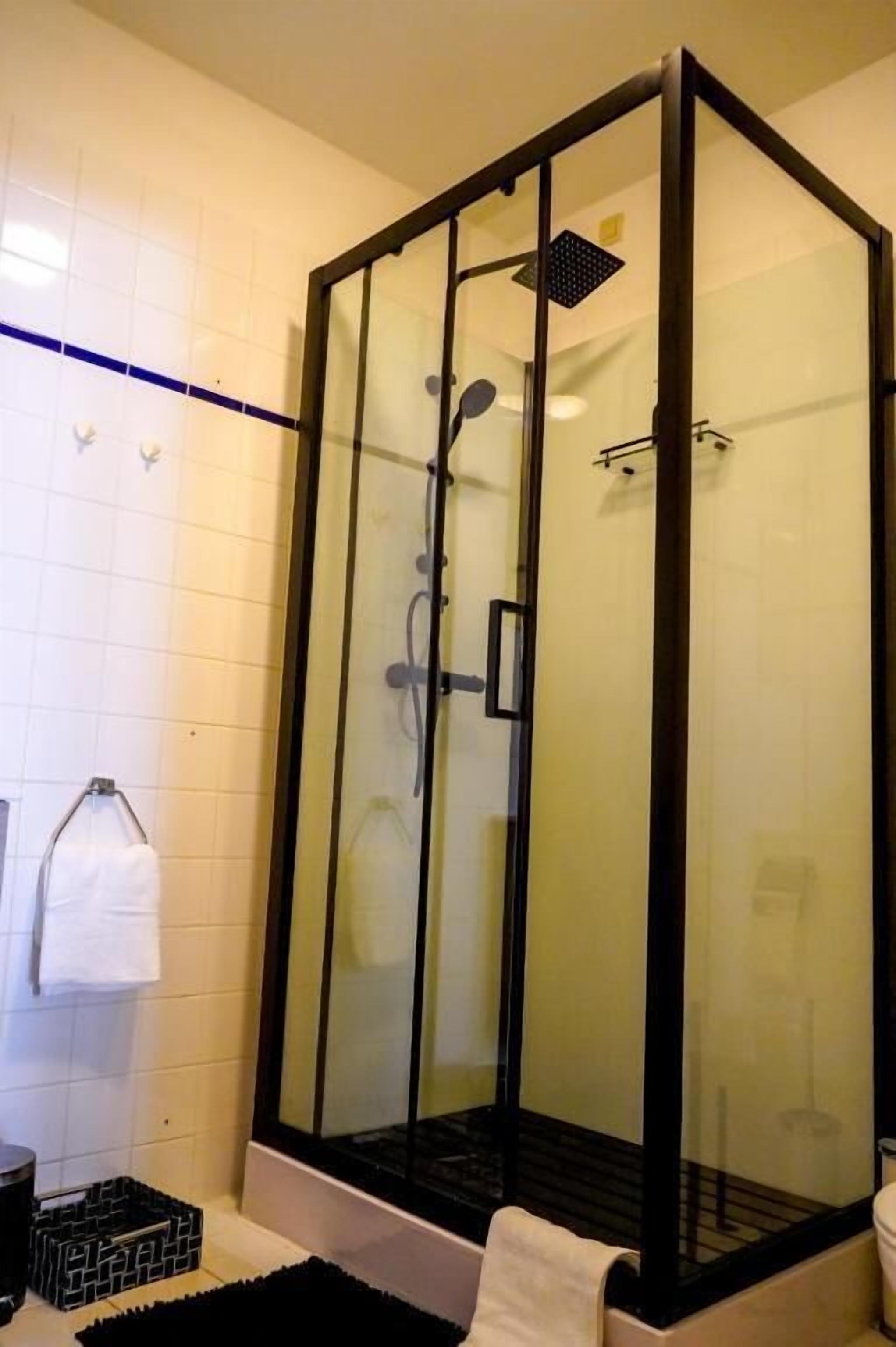 Une cabine de douche fermée par des parois en verre, avec un cadre noir, un mur recouvert de carreaux blancs et un tapis noir au sol.