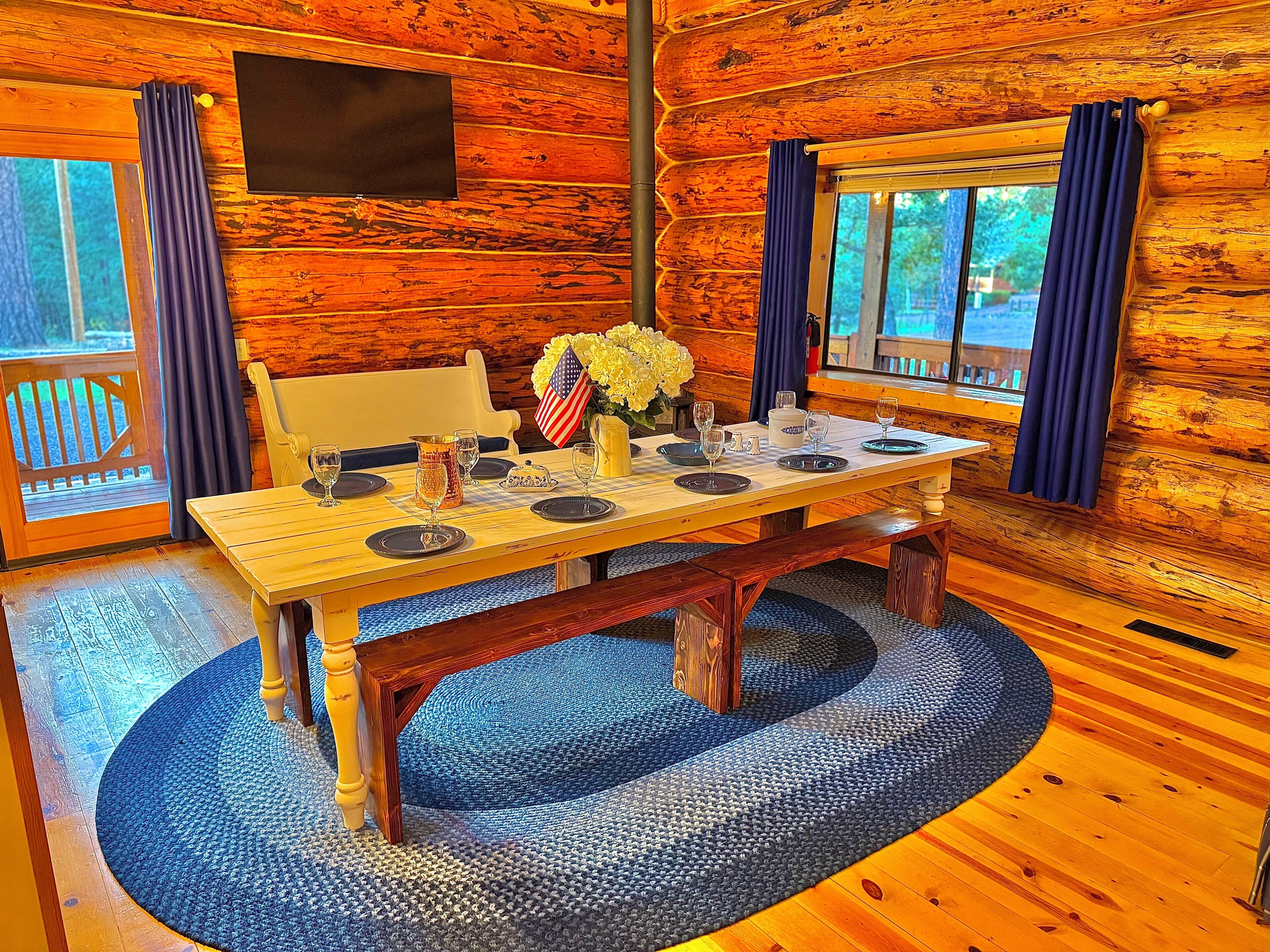 Interior de una cabaña de madera con una mesa de comedor dispuesta para cuatro personas, una bandera y un jarrón con flores.