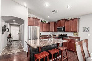 A modern kitchen with granite countertops, stainless steel appliances, wooden cabinets, and a bar area with stools. - A Maricopa Must: 4BR/3BA Immaculate Home w/Jacuzzi (Maricopa)