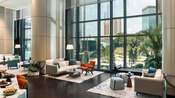 Eine geräumige Lobby mit einem großen Fenster und Blick auf die Stadt, eine Sitzecke mit Sofas und Sesseln sowie ein Couchtisch mit Snacks.