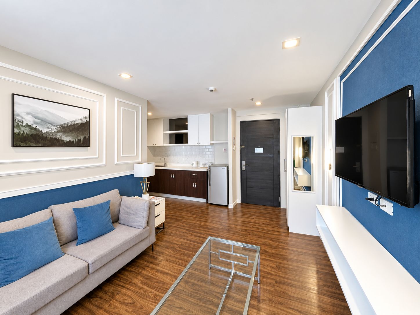 A modern living room with a sofa, a flat-screen TV, and a kitchen in the background.