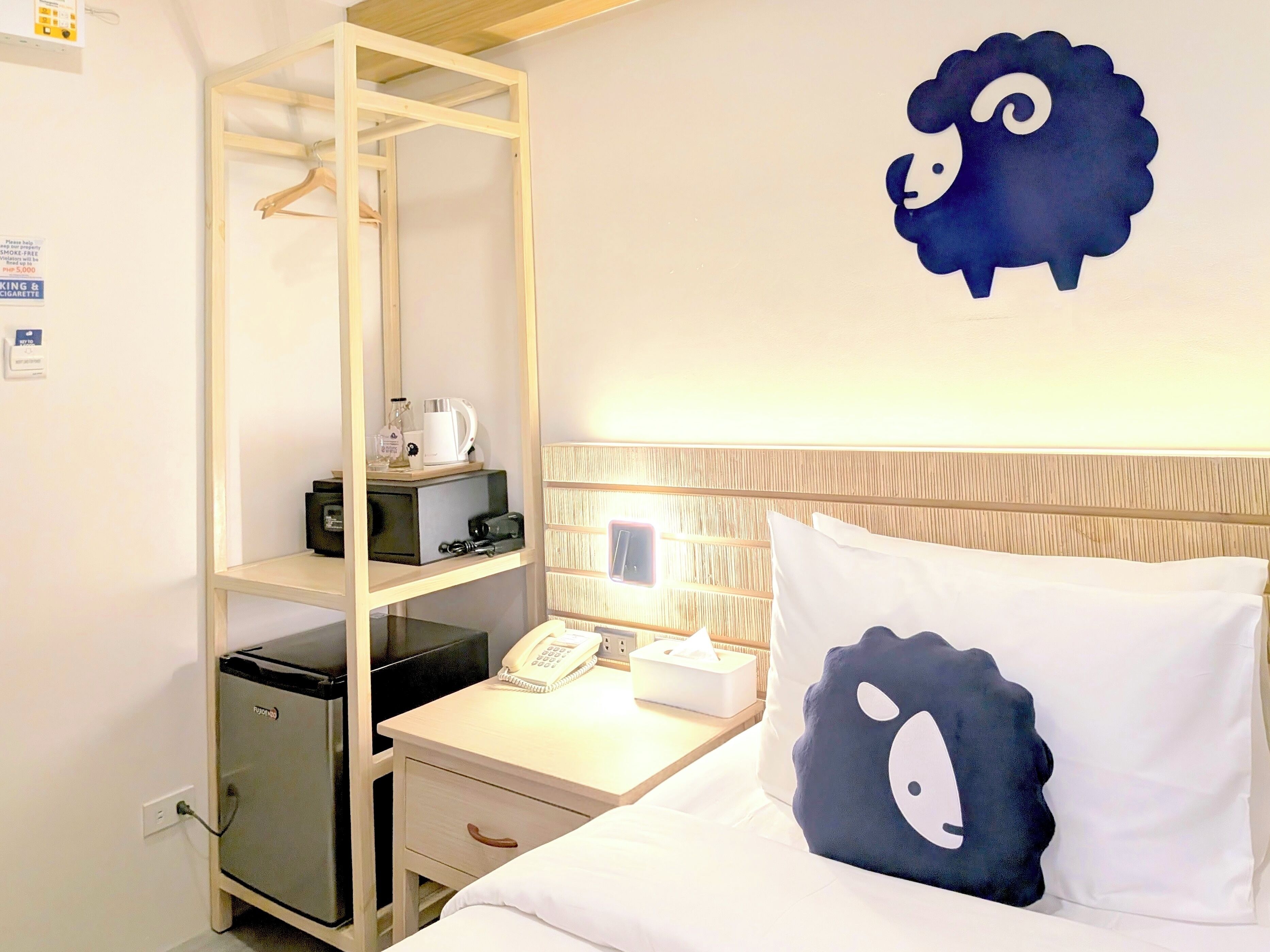 Une chambre d’hôtel comprenant un lit, une table de chevet avec une lampe, un petit bureau avec un téléphone et une boîte de mouchoirs, une armoire et un élément décoratif mural représentant un mouton.