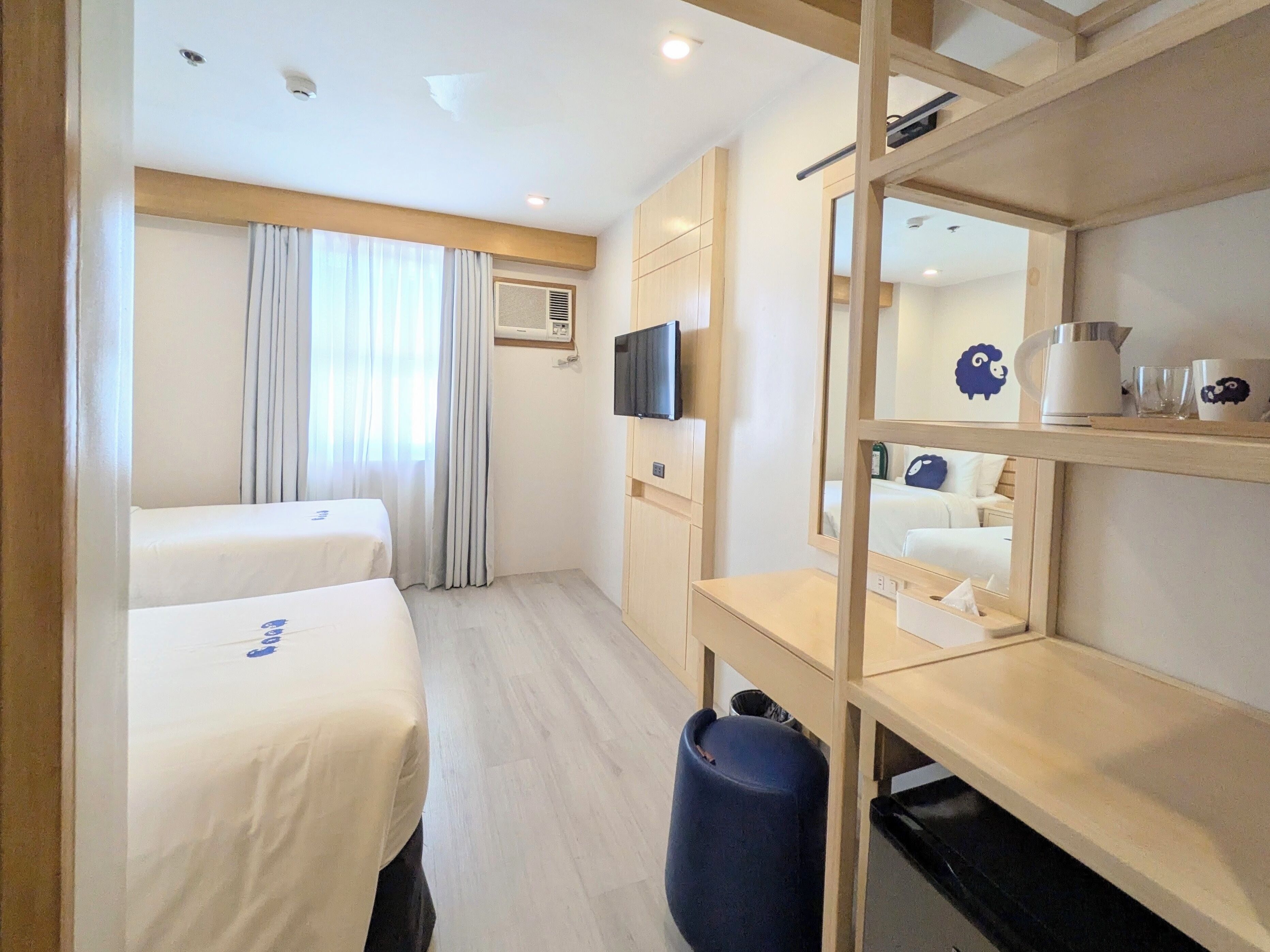 A hotel room with two beds, a TV, a mirror, a shelf with items, and a trash bin.