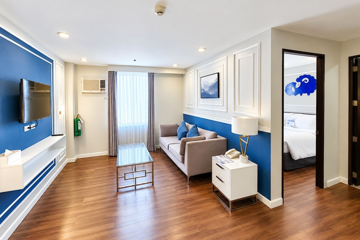 Quarto de hotel moderno com televisão de tela plana, sofá, mesa pequena e luminária de cabeceira.