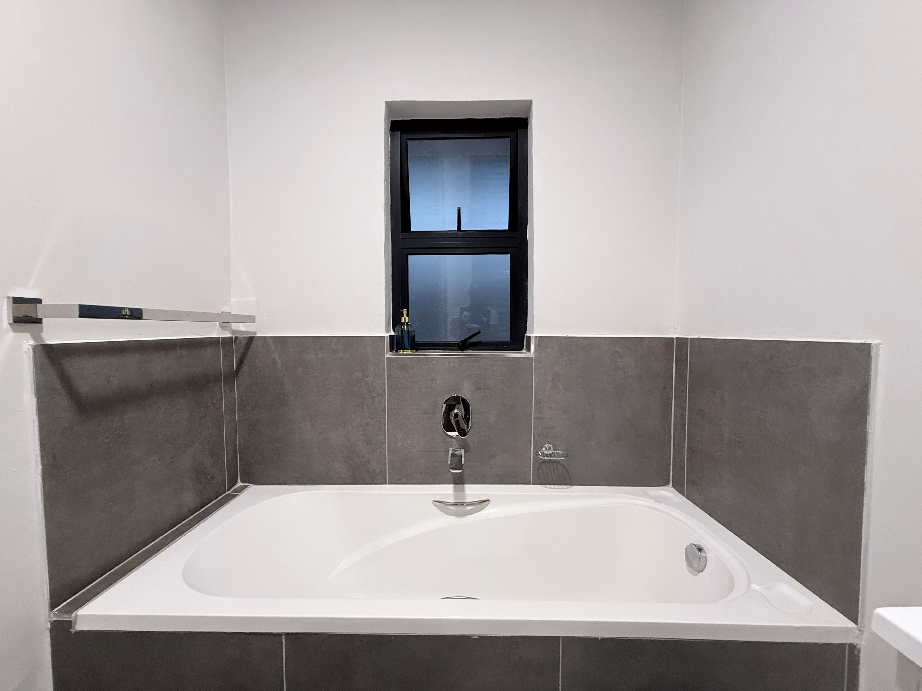 Et badeværelse med badekar, et vindue og grå fliserede vægge.