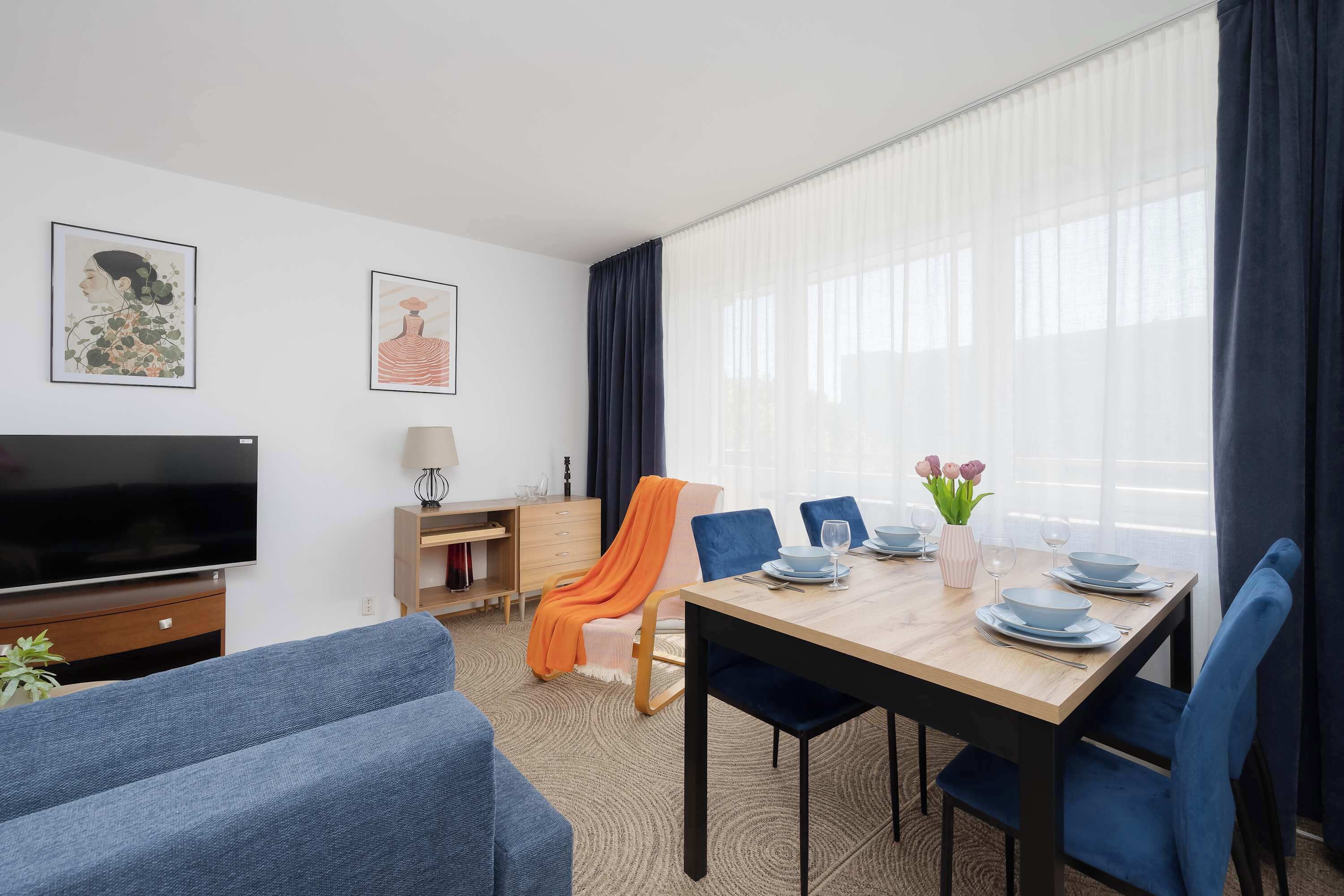 Ett modernt rum med en platt-TV, ett matbord dukat med tallrikar och glas, en soffa och ett skrivbord med en lampa.