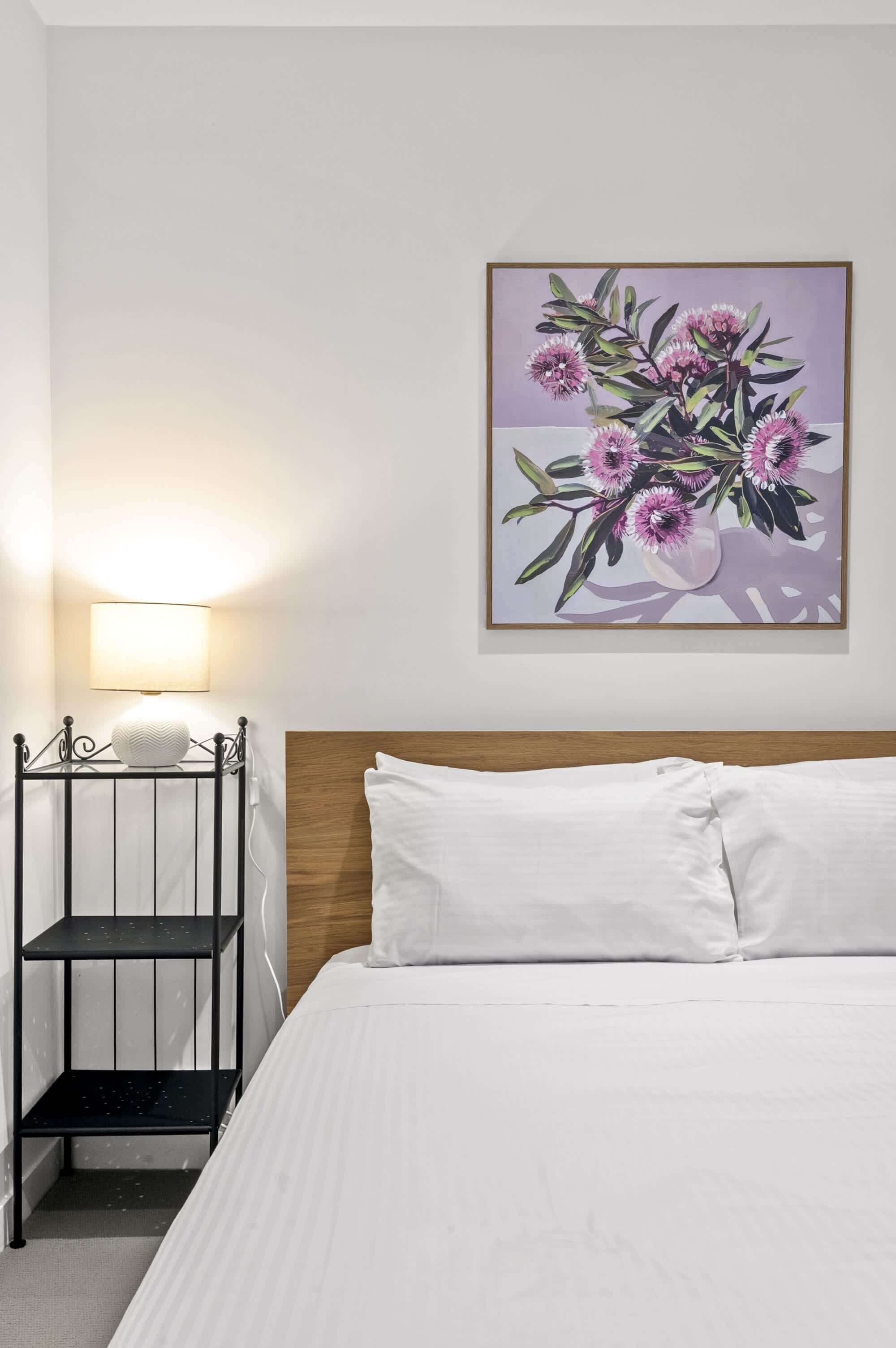 Ein Hotelzimmer mit einem Bett, einem Nachttisch mit Lampe, einem Regal und einem gerahmten Blumenbild an der Wand.