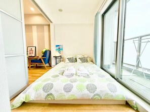 Ein schmales Zimmer mit einem Bett, einem Sessel, einem kleinen Tisch und einem Fenster mit Blick auf eine Brücke.