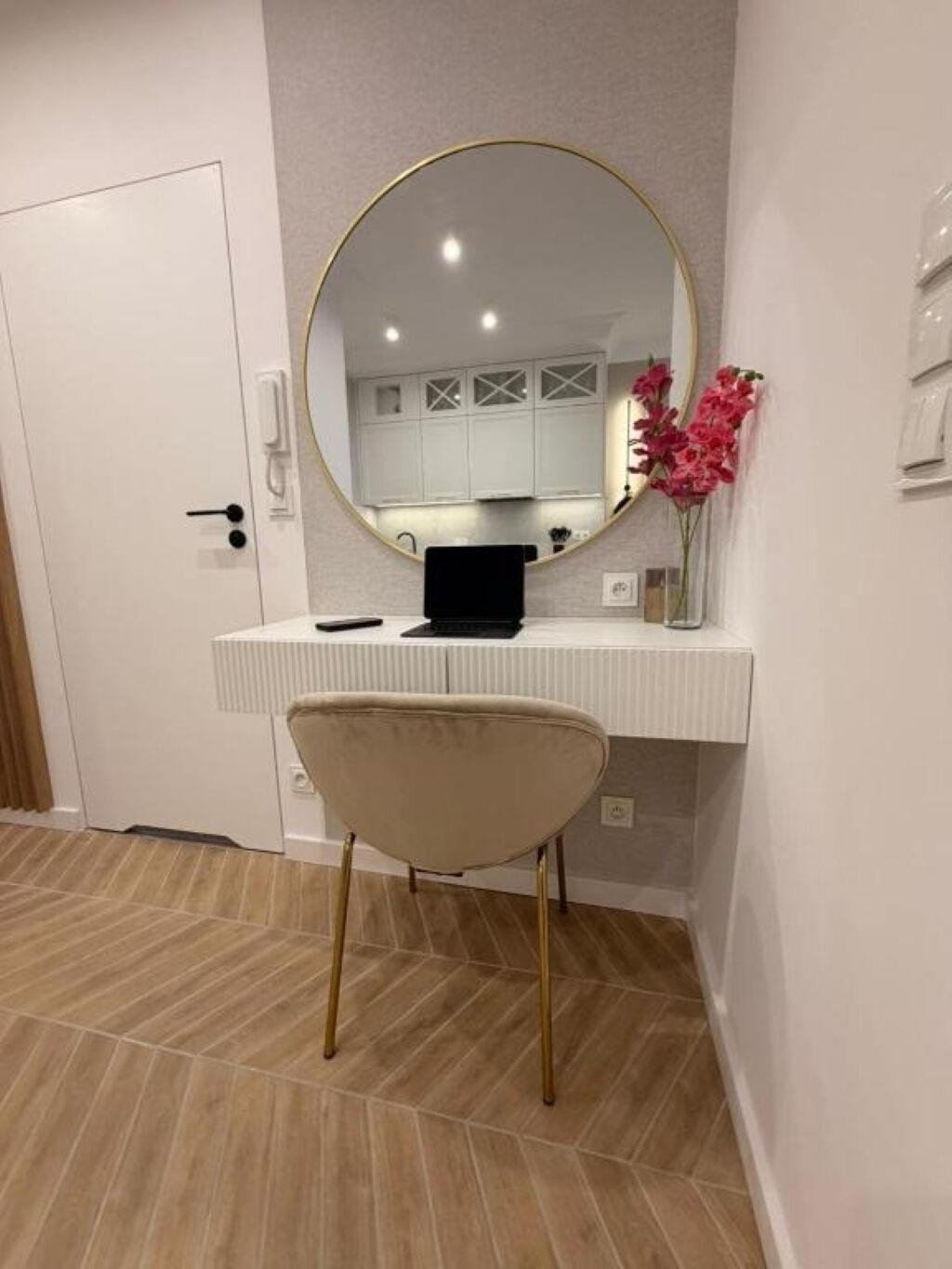 Una habitación con una puerta blanca, un espejo redondo sobre un escritorio blanco, una laptop en el escritorio, una silla y un florero con flores.