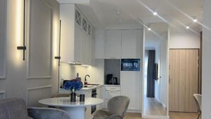 A modern living room with a grey sofa, a small dining table, and a kitchen area with white cabinets and a blue appliance.