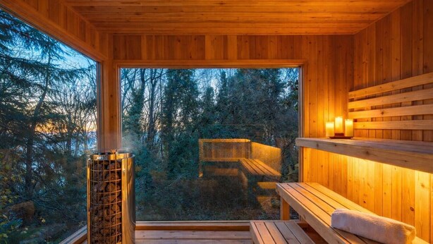 Een houten sauna met een bank en een sauna-kachel, een raam met uitzicht op bomen, en een plankje met kaarsen.