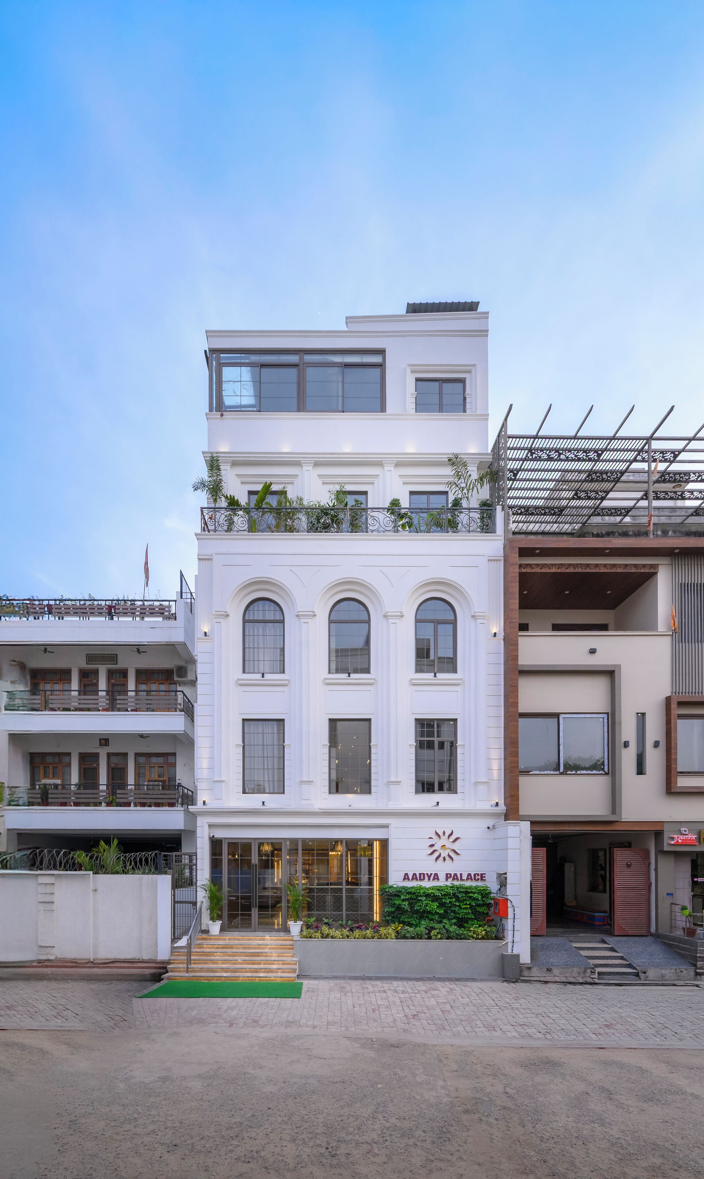 Un edificio de varios pisos con una entrada destacada, un balcón con plantas y un letrero que dice 'AADYA PALACE'.