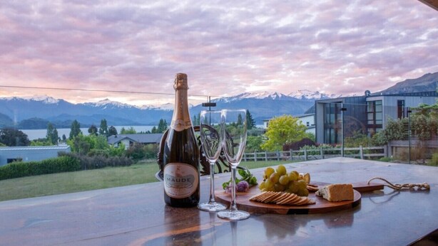 Une bouteille de vin, un verre, des raisins et des crackers sur une table, avec vue sur des montagnes et un lac.