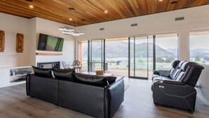 Un salon moderne avec un canapé en cuir noir, un fauteuil inclinable en cuir noir, un téléviseur à écran plat et une grande fenêtre offrant une vue sur les montagnes et un lac.
