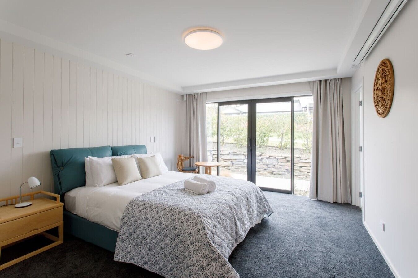 A bedroom with a bed, bedside table, a small table, and a view of the outdoors.