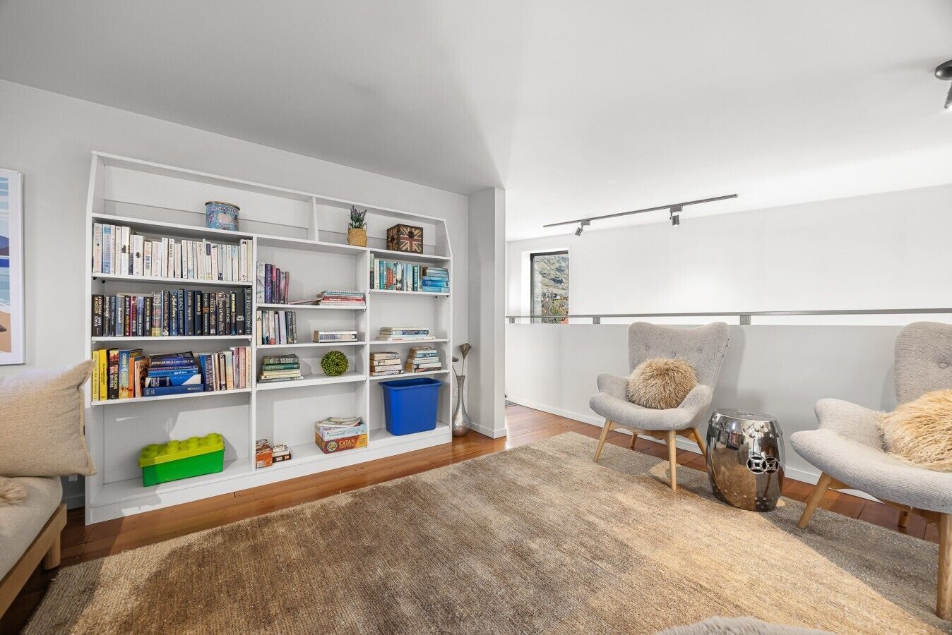 A modern living room with a white bookshelf, two grey armchairs, a wooden floor, and a large window.