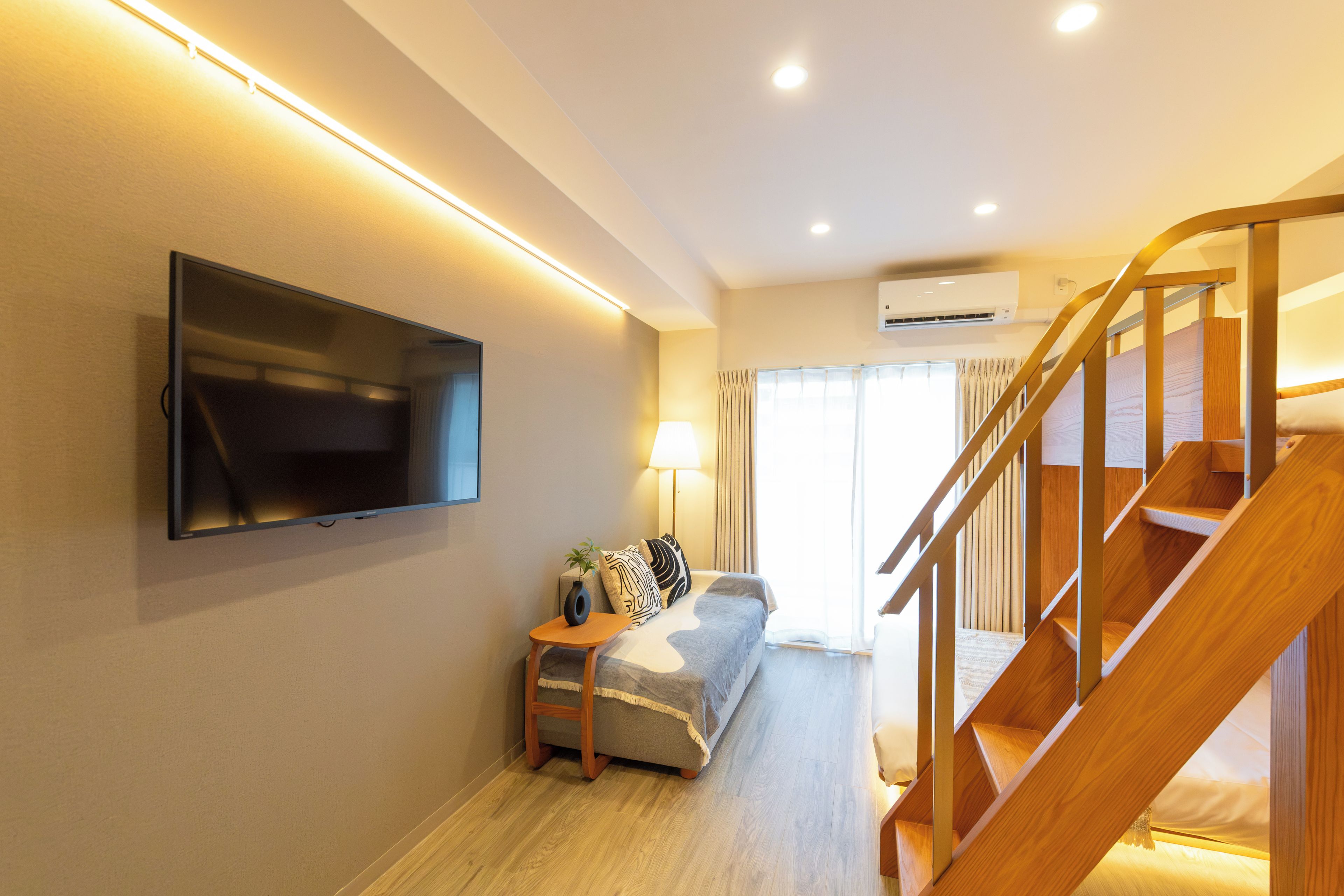 A modern living room with a flat-screen TV mounted on the wall, a sofa with patterned cushions, a small wooden side table, and a staircase with wooden railings.