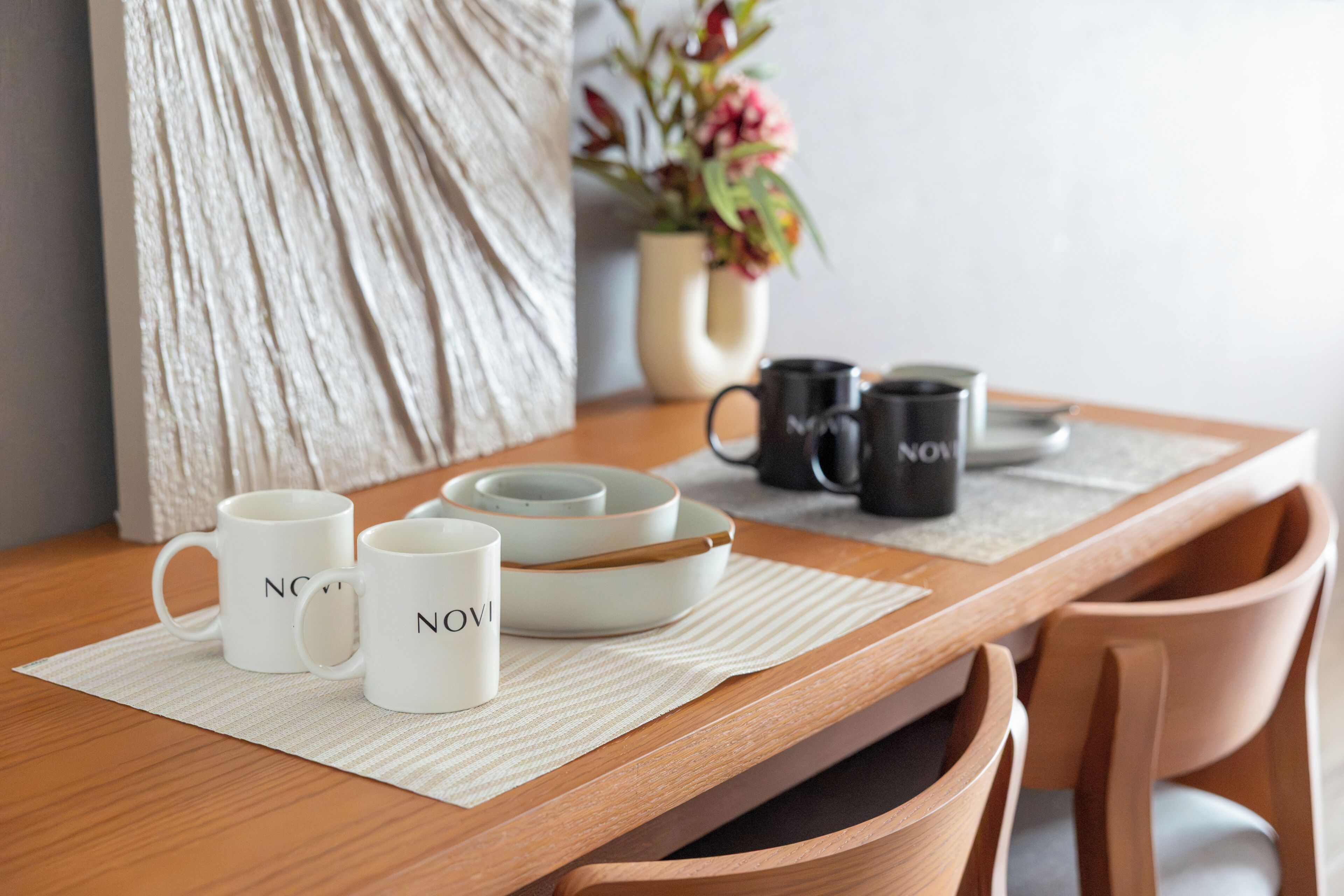 Une table en bois, avec des tasses et des assiettes en céramique blanche, un set de table en fibres tressées et un vase rempli de fleurs.