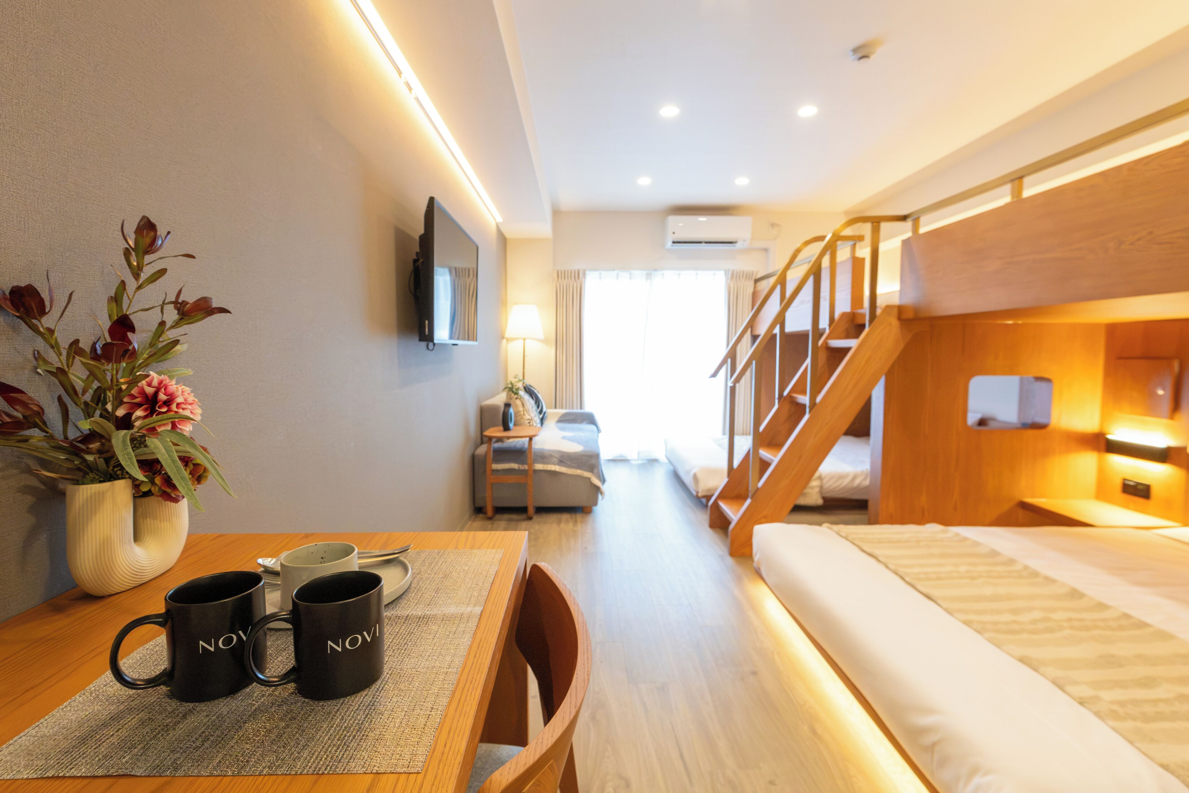 A hotel room with a bunk bed, a desk with a vase of flowers, and a table with two mugs.