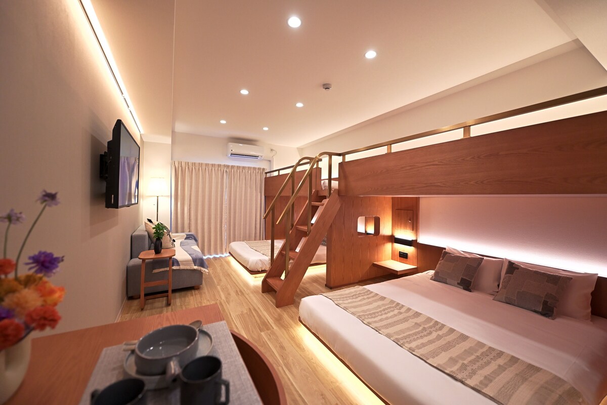 A hotel room with a bunk bed, a desk with a chair, a small table with a plant, and a window with curtains.